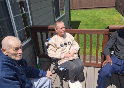 Three people are sitting outside on a wooden deck in the sun, engaging in conversation. The background shows a grassy yard and neighboring buildings.