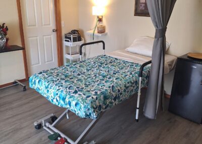A hospital room featuring a single bed with floral bedding, a bedside table with a lamp, and a privacy curtain. The room includes a door, a clock, and minimal decor.