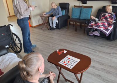 A group of people in a living room engage in different activities such as watching TV, playing games, and conversing. A wheelchair and some balloons are in the background.